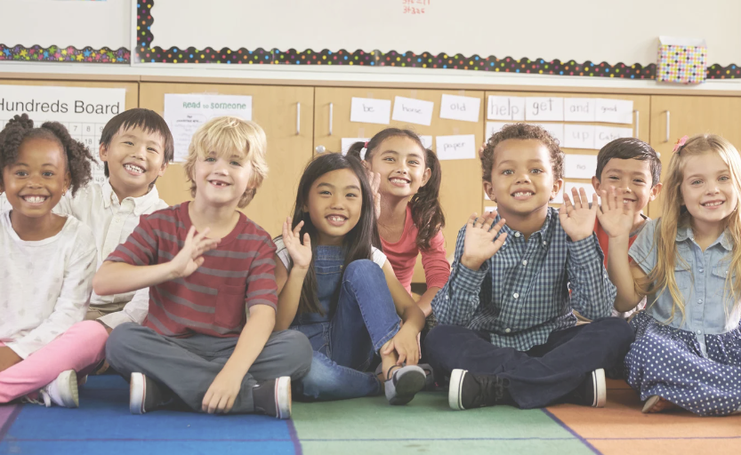 diverse group of children sitting in a classroom waving